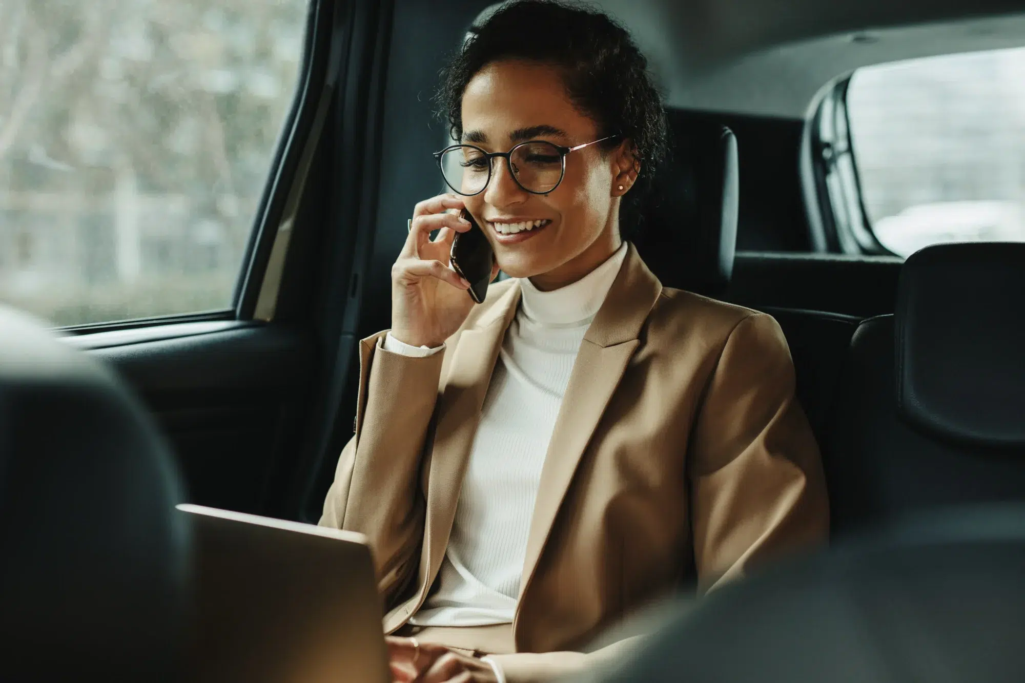 Woman sitting in the back of the car talking on the phone while working on her laptop